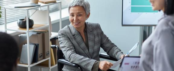 A woman using a wheelchair and wearing a grey business suit meets with work colleagues.