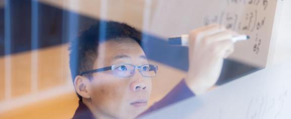 Georgia Tech Assistant Professor Glen Chou with the School of Cybersecurity and Privacy works through an equation on a transparent writing board.